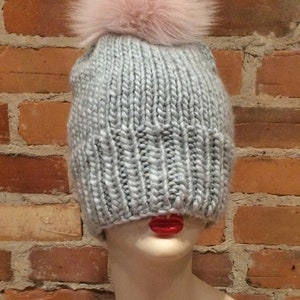 May include: A light gray knit beanie with a large, fluffy, light pink pom-pom on top. The hat is displayed on a mannequin head with red lips, set against a brick wall background.