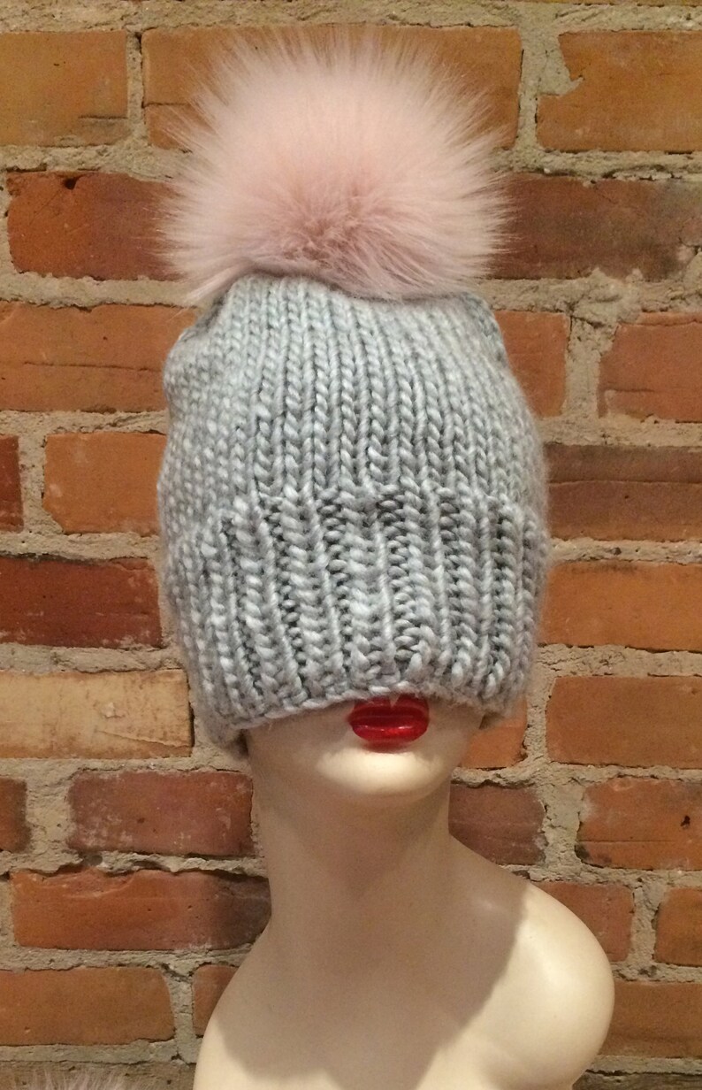 May include: A light gray knit beanie with a large, fluffy, light pink pom-pom on top. The hat is displayed on a mannequin head with red lips, set against a brick wall background.