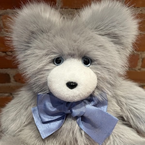 K&ouml;nnte beinhalten: Ein grauer Pl&uuml;schteddyb&auml;r mit wei&szlig;em Gesicht und blauer Schleife. Der B&auml;r hat blaue Augen und eine schwarze Nase.