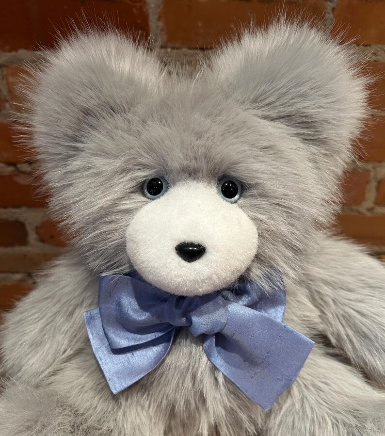 K&ouml;nnte beinhalten: Ein grauer Pl&uuml;schteddyb&auml;r mit wei&szlig;em Gesicht und blauer Schleife. Der B&auml;r hat blaue Augen und eine schwarze Nase.