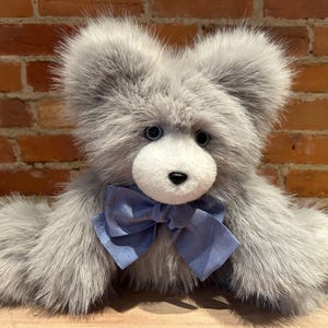 K&ouml;nnte beinhalten: Ein grauer Pl&uuml;schteddyb&auml;r mit einer blauen Schleife. Der B&auml;r hat ein wei&szlig;es Gesicht mit schwarzen Augen und einer schwarzen Nase.