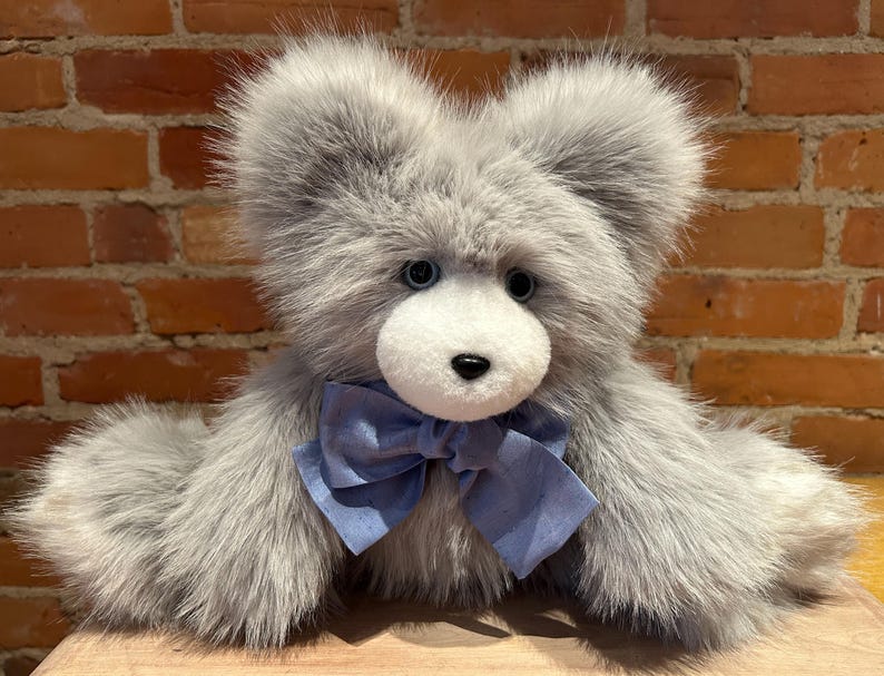 K&ouml;nnte beinhalten: Ein grauer Pl&uuml;schteddyb&auml;r mit einer blauen Schleife. Der B&auml;r hat ein wei&szlig;es Gesicht mit schwarzen Augen und einer schwarzen Nase.