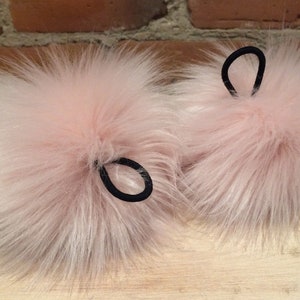 May include: Two fluffy, light pink faux fur pom-poms with black elastic hair ties. The pom-poms have long, soft fibers and are likely used as accessories. The background is a blurred wooden surface and a brick wall.