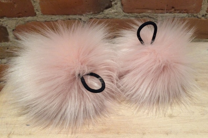 May include: Two fluffy, light pink faux fur pom-poms with black elastic hair ties. The pom-poms have long, soft fibers and are likely used as accessories. The background is a blurred wooden surface and a brick wall.