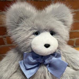 K&ouml;nnte beinhalten: Ein pl&uuml;schiger grauer Teddyb&auml;r mit wei&szlig;em Gesicht und blauen Augen. Der B&auml;r tr&auml;gt eine hellblaue Schleife aus Satin.