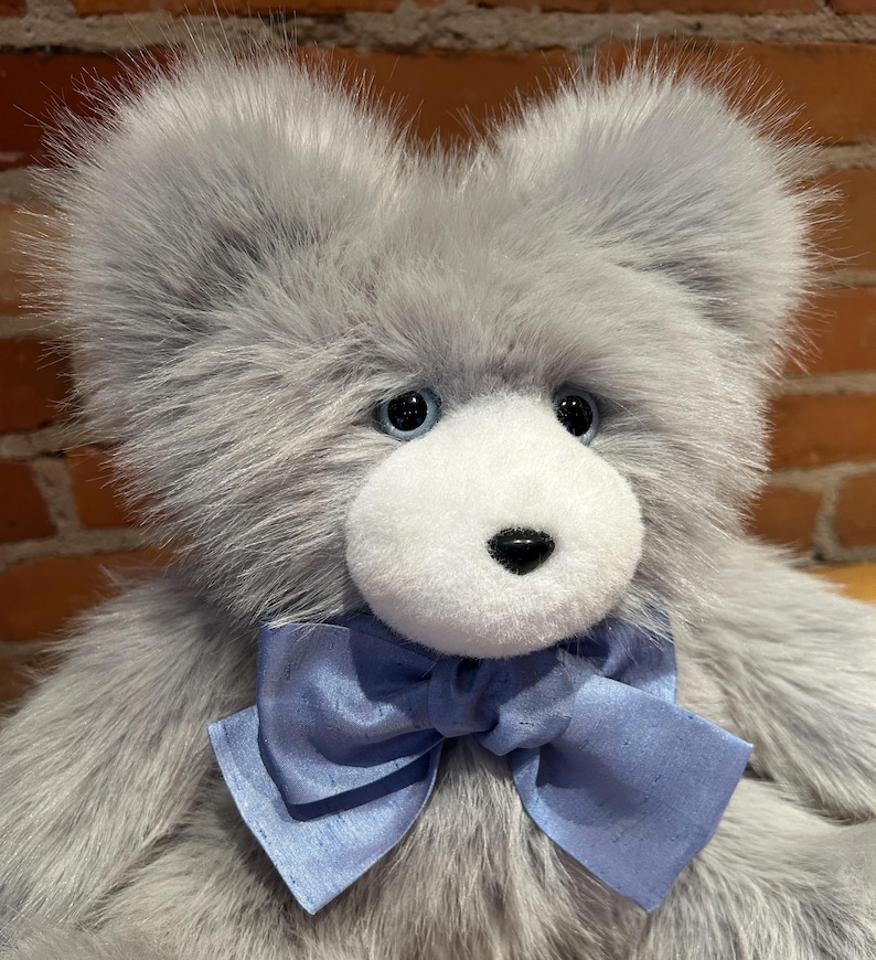 K&ouml;nnte beinhalten: Ein pl&uuml;schiger grauer Teddyb&auml;r mit wei&szlig;em Gesicht und blauen Augen. Der B&auml;r tr&auml;gt eine hellblaue Schleife aus Satin.
