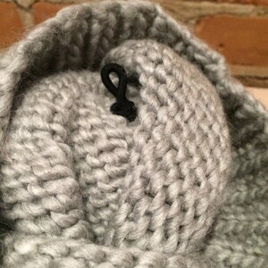 May include: Close-up of a gray knitted hat with a black cord detail. The hat is made of thick, chunky yarn, and the texture of the knit is clearly visible. The background is a blurred brick wall.