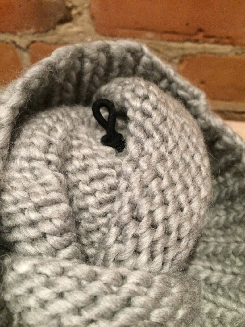 May include: Close-up of a gray knitted hat with a black cord detail. The hat is made of thick, chunky yarn, and the texture of the knit is clearly visible. The background is a blurred brick wall.