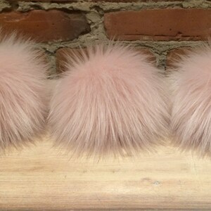 May include: Three fluffy, light pink fur pom-poms. The pom-poms are round and have a dense, textured appearance. They are arranged in a row on a wooden surface, with a brick wall in the background.