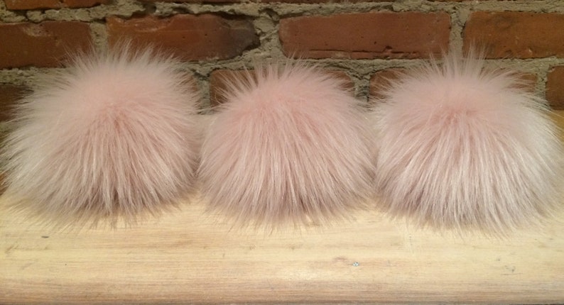 May include: Three fluffy, light pink fur pom-poms. The pom-poms are round and have a dense, textured appearance. They are arranged in a row on a wooden surface, with a brick wall in the background.
