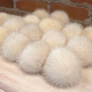 Peut inclure: Un ensemble de pompons duveteux de couleur crème disposés sur une surface en bois clair. Les pompons ont un aspect doux et texturé et sont regroupés. Un mur de briques est visible en arrière-plan.