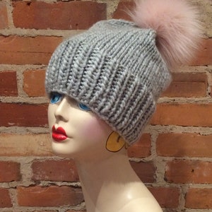 May include: A gray knit beanie with a large pink fur pom-pom. The hat is made of thick, ribbed yarn. The beanie is displayed on a mannequin head with red lipstick and blue eyes. The background is a brick wall.
