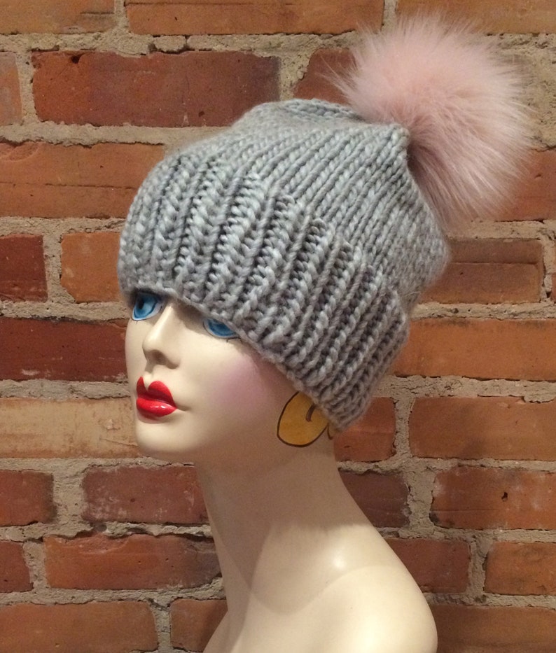 May include: A gray knit beanie with a large pink fur pom-pom. The hat is made of thick, ribbed yarn. The beanie is displayed on a mannequin head with red lipstick and blue eyes. The background is a brick wall.
