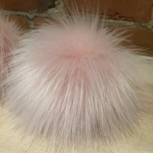 May include: A fluffy, light pink pompom with long, soft, and textured fibers. The pompom is round and appears to be made of faux fur. The background includes a brick wall and a wooden surface.