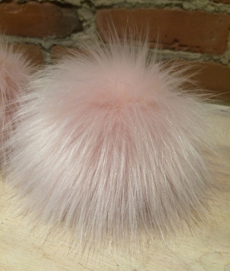 May include: A fluffy, light pink pompom with long, soft, and textured fibers. The pompom is round and appears to be made of faux fur. The background includes a brick wall and a wooden surface.