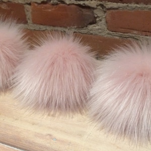 May include: Three fluffy, light pink faux fur pom poms. The pom poms are round and have long, soft, and wispy fibers. They are arranged in a row on a light-colored wooden surface, with a brick wall in the background.
