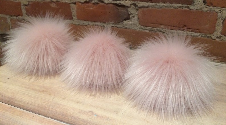 May include: Three fluffy, light pink faux fur pom poms. The pom poms are round and have long, soft, and wispy fibers. They are arranged in a row on a light-colored wooden surface, with a brick wall in the background.