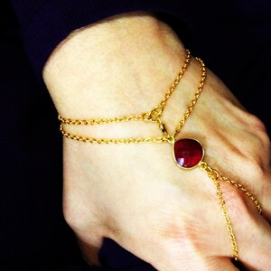 May include: A gold chain bracelet with a red gemstone pendant. The bracelet is double-layered and has a finger loop.
