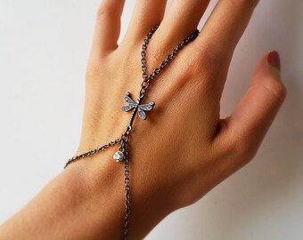 May include: A hand-chain bracelet featuring a dragonfly charm. The bracelet is made of dark metal and has a dragonfly pendant with small, clear stones. A small, round, clear stone hangs below the dragonfly.