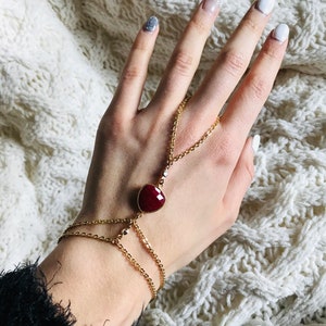 Peut inclure: Un bracelet en chaîne dorée avec un pendentif en forme de cœur en pierre rouge. Le bracelet est conçu pour être porté au poignet et au doigt, avec une chaîne reliant les deux.