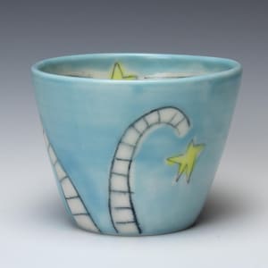 May include: A light blue ceramic cup with a whimsical design. The cup features a curved, segmented line pattern and yellow star accents. The cup's exterior is a solid light blue color, and the interior is a darker blue.