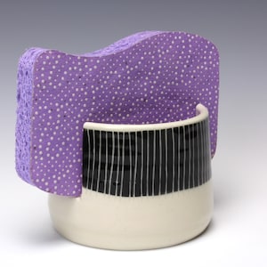 May include: A ceramic sponge holder with a purple, dotted sponge insert. The holder has a black and white striped design on the upper portion and a white base. The sponge insert is a light purple color with white dots.