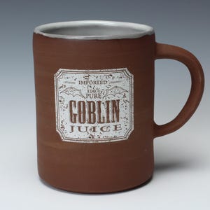 May include: A brown ceramic mug with a handle and a white rim. The mug features a rectangular label with the words "GOBLIN JUICE" in a distressed font. The label also includes the text "IMPORTED" and "100% PURE".