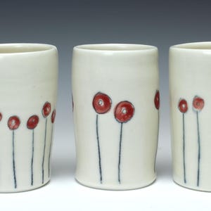 May include: Three white ceramic cups with red flower designs. The cups are all the same size and shape, and they have a simple, elegant design.
