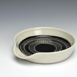 May include: A white ceramic spoon rest with a black center featuring a concentric circular design. The spoon rest has a curved shape with a small lip on one side. The design is a modern, minimalist style.