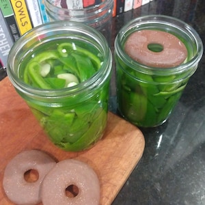 May include: Two glass jars filled with green peppers and a brown disc on top of the peppers in one jar. Two brown discs with a hole in the center are on a wooden cutting board.