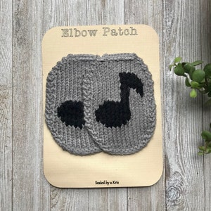 Può includere: Due toppe per gomito in maglia grigia con disegni di note musicali nere. Le toppe sono esposte su un pannello di legno con il testo "Elbow Patch" e "Sealed by a Kris".