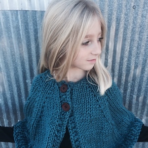 Peut inclure: Une cape en tricot bleu sarcelle avec des boutons marrons. La cape a un col rond et une base légèrement évasée.