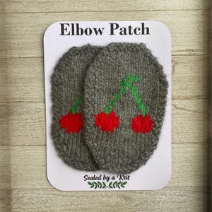 Puede incluir: Dos parches de codo de punto gris con cerezas rojas y tallos verdes. Los parches están en una tarjeta blanca con el texto "Elbow Patch" y "Sealed by a Kris".