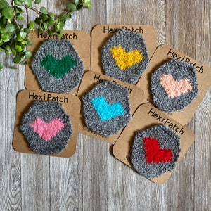 May include: Six grey hexagon-shaped coasters with a knitted heart design in different colours: green, yellow, pink, blue, red, and pink. Each coaster has a brown paper backing with the text "HexiPatch" printed on it.