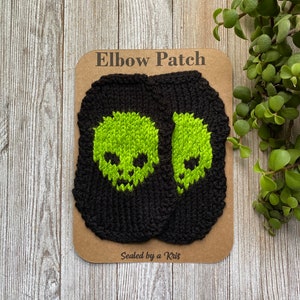 May include: Two black knitted elbow patches with green skull designs. The patches are packaged on a brown card with the text "Elbow Patch" and "Sealed by a Kris".