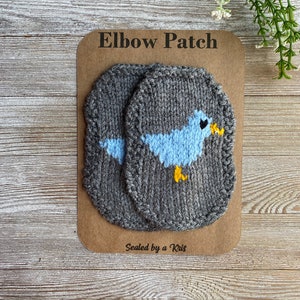 May include: Two gray knit elbow patches with a light blue bird design and yellow beak and feet. The patches are on a brown card with the text "Elbow Patch" and "Sealed by a Kris".