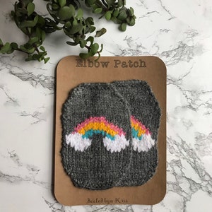 Puede incluir: Dos parches grises para codos con diseños de arcoíris. Cada parche presenta un arcoíris con colores rosa, amarillo, azul y blanco. Los parches se muestran en una tarjeta marrón con el texto "Elbow Patch" y "Sealed by a Kris".