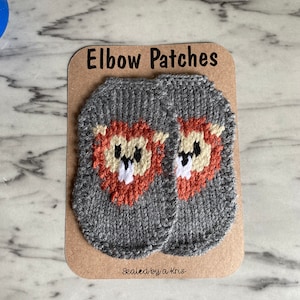 Puede incluir: Dos parches de codo de punto gris con caras de león naranja. Los parches están en una tarjeta marrón con el texto "Elbow Patches" y "Sealed by a Kris".