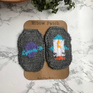 May include: Two gray knitted elbow patches with embroidered designs. One patch features a purple planet with a blue ring, and the other features a white rocket with orange and blue details. The patches are on a brown card with the text "Elbow Patch" and "Sealed by a Kris".