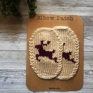 Puede incluir: Dos parches de codo de punto beige con un diseño de reno burdeos. Los parches están en una tarjeta marrón con el texto "Elbow Patch" y "Sealed by a Kris".