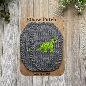 May include: Gray knitted elbow patch with a lime green dinosaur design. The patch is on a brown card with the words "Elbow Patch" and "Sealed by a Kris". The patch is a unique accessory for clothing repair or customization.