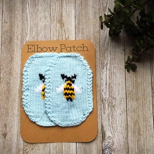 May include: Light blue knitted elbow patches with embroidered bee designs. The patches are displayed on a brown card with the text "Elbow Patch". The bees have black and yellow stripes and white wings.