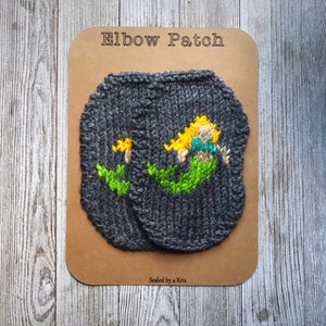 Puede incluir: Un par de parches de codo de punto gris con un diseño de sirena bordado en un parche. La sirena es amarilla, verde y azul. Los parches están en una tarjeta marrón con el texto "Elbow Patch" y "Sewed by a Kris".