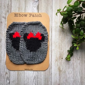 Può includere: Toppe per gomiti in maglia grigia con disegni neri e rossi a forma di topo. Le toppe sono esposte su una carta marrone con la scritta "Elbow Patch". Lo sfondo è una superficie di legno chiaro con fogliame verde.