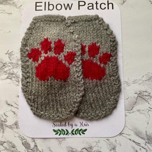 Puede incluir: Dos parches de codo grises tejidos con huellas de patas rojas. Los parches están sobre una tarjeta blanca con el texto "Elbow Patch" en la parte superior y "Sealed by a Kris" en la parte inferior. El fondo es un patrón de mármol.