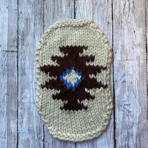 May include: Cream-colored, hand-knitted coaster with a brown and blue geometric design. The coaster has a rounded rectangular shape and is made of thick yarn. The design is centered on the coaster and features a symmetrical pattern.