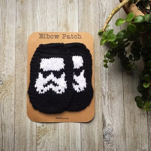 May include: Two black and white knitted elbow patches with a Star Wars Stormtrooper design. The patches are on a brown card with the text "Elbow Patch" and "Sealed by a Kris".