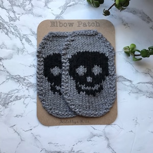 Könnte beinhalten: Zwei graue gestrickte Ellenbogen-Patches mit schwarzen Totenkopf-Designs. Die Patches befinden sich auf einer braunen Karte mit dem Text "Elbow Patch" und "Sealed by a Kris".