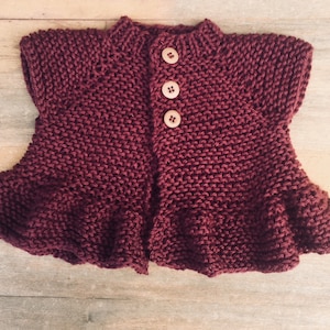 May include: A maroon knitted cardigan with a button closure and a ruffled hem.