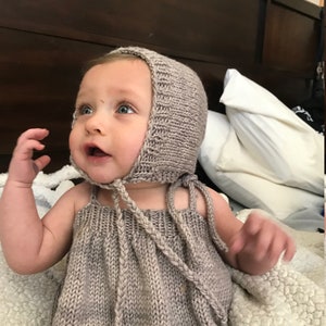 May include: A baby wearing a knitted hooded dress and matching bonnet. The dress and bonnet are a light brown color with a subtle marbled pattern.